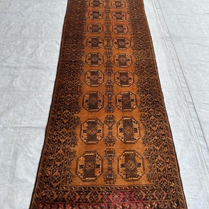 May include: A long, rectangular runner rug featuring a warm, burnt orange background with repeating dark brown geometric octagonal motifs. The rug is framed by a detailed border with intricate patterns and finished with short, grey fringe at both ends.