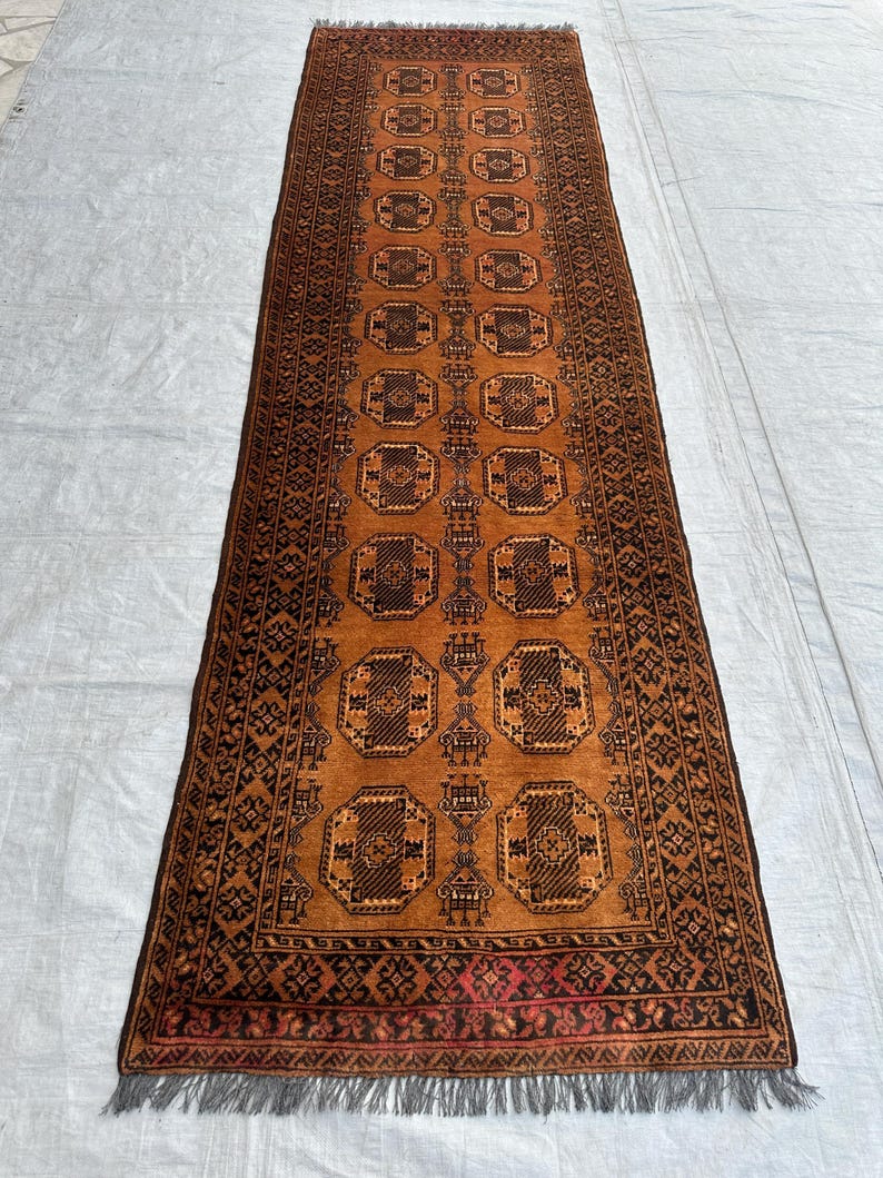 May include: A long, rectangular runner rug featuring a warm, burnt orange background with repeating dark brown geometric octagonal motifs. The rug is framed by a detailed border with intricate patterns and finished with short, grey fringe at both ends.
