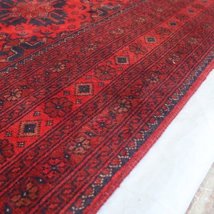 May include: Close-up of a red rug with a black and brown geometric pattern. The rug has a fringe border and is folded over on itself.