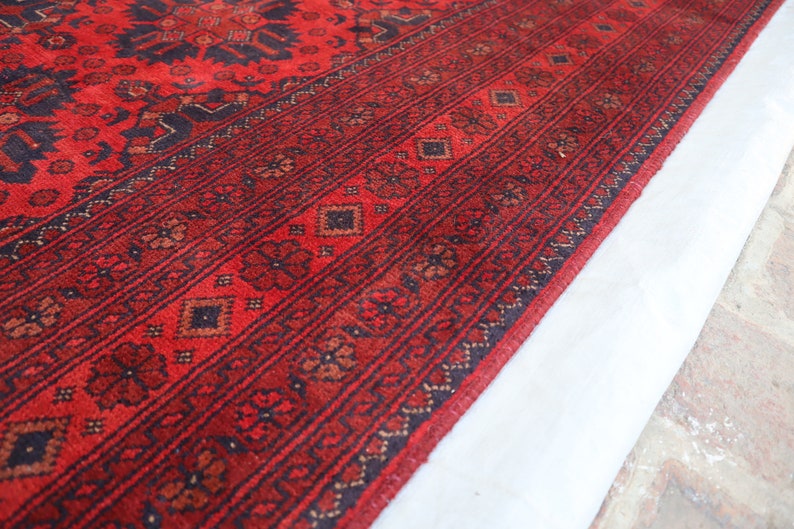 May include: Close-up of a red rug with a black and brown geometric pattern. The rug has a fringe border and is folded over on itself.
