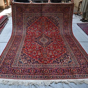 Puede incluir: Una alfombra rectangular grande que presenta un campo rojo vibrante con un medall&oacute;n central azul marino y patrones florales intrincados en tonos azul, crema y dorado. La alfombra tiene un borde decorativo ancho y flecos blancos abajo.