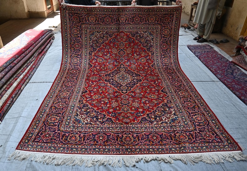 Puede incluir: Una alfombra rectangular grande que presenta un campo rojo vibrante con un medall&oacute;n central azul marino y patrones florales intrincados en tonos azul, crema y dorado. La alfombra tiene un borde decorativo ancho y flecos blancos abajo.