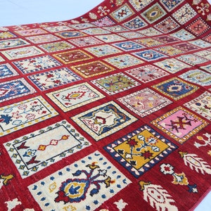 May include: A large, rectangular red rug with a repeating pattern of colorful square designs. Each square contains a unique floral or geometric motif in shades of blue, yellow, pink, and white. The rug's border features stylized floral elements.