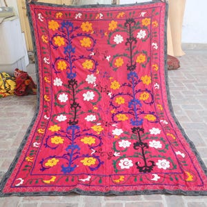 K&ouml;nnte beinhalten: Rechteckiger Textil mit tiefrosa Hintergrund. Das Design zeigt stilisierte Blumenmuster in Gelb, Wei&szlig;, Blau und Gr&uuml;n, umrahmt von einem dunklen Rand. Das Textil wird wahrscheinlich als dekorativer Wandbehang oder &Uuml;berwurf verwendet.