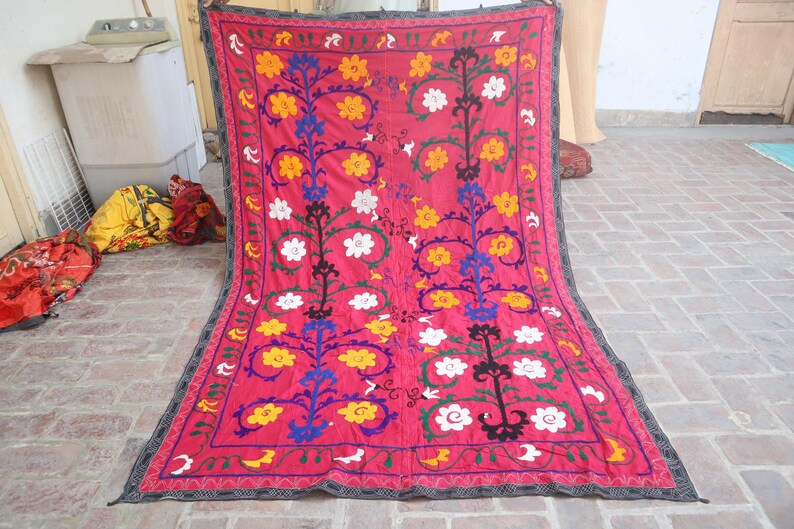 K&ouml;nnte beinhalten: Rechteckiger Textil mit tiefrosa Hintergrund. Das Design zeigt stilisierte Blumenmuster in Gelb, Wei&szlig;, Blau und Gr&uuml;n, umrahmt von einem dunklen Rand. Das Textil wird wahrscheinlich als dekorativer Wandbehang oder &Uuml;berwurf verwendet.