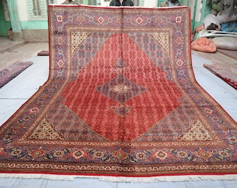 May include: A large, rectangular red rug with an intricate geometric pattern. The rug features a central diamond design in a darker shade of red, surrounded by a border of floral and geometric motifs in shades of blue, beige, and brown. The rug is displayed on a light-coloured surface.