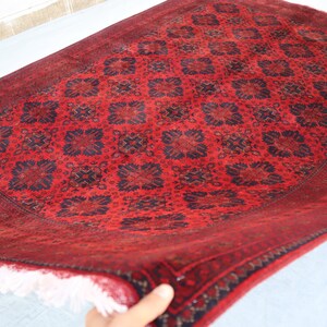 May include: A red and blue patterned rug with a white fringe. The rug is folded in half and is being held by a hand.