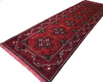 Puede incluir: Una alfombra pasillera rectangular larga con un fondo rojo intenso y motivos geométricos de diamantes en azul oscuro y negro. La alfombra está decorada con intrincados patrones en blanco y tonos dorados, con flecos cortos en ambos extremos.