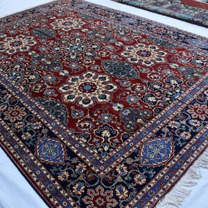 K&ouml;nnte beinhalten: Ein rechteckiger Teppich mit einem tiefroten Mittelfeld, das komplexe Blumen- und Medaillonmuster in Marineblau, Creme und Hellblau aufweist. Der Teppich hat eine breite marineblaue Bord&uuml;re mit wiederkehrenden Blumenmotiven und kurze wei&szlig;e Fransen.