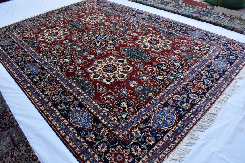 K&ouml;nnte beinhalten: Ein rechteckiger Teppich mit einem tiefroten Mittelfeld, das komplexe Blumen- und Medaillonmuster in Marineblau, Creme und Hellblau aufweist. Der Teppich hat eine breite marineblaue Bord&uuml;re mit wiederkehrenden Blumenmotiven und kurze wei&szlig;e Fransen.