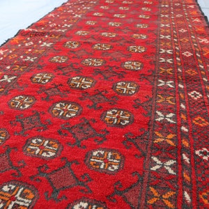 May include: A red rug with a geometric pattern in black, orange, and white. The rug has a border with a repeating design.