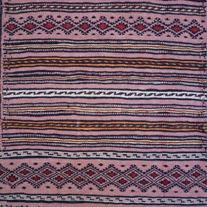 May include: A woven textile featuring a dusty pink base with a central rectangular panel of horizontal stripes in black, white, and orange. The border displays a repeating geometric diamond pattern in dark blue and red, framing the design.