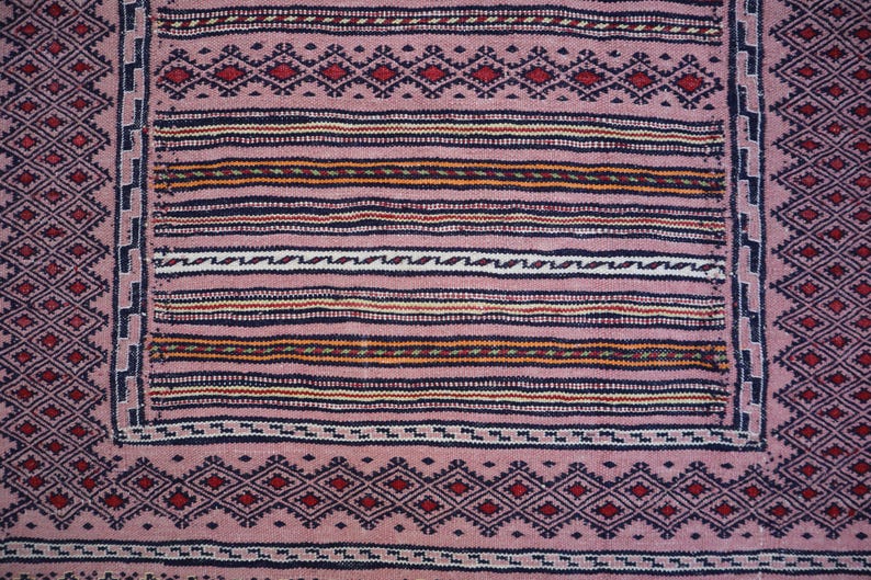 May include: A woven textile featuring a dusty pink base with a central rectangular panel of horizontal stripes in black, white, and orange. The border displays a repeating geometric diamond pattern in dark blue and red, framing the design.