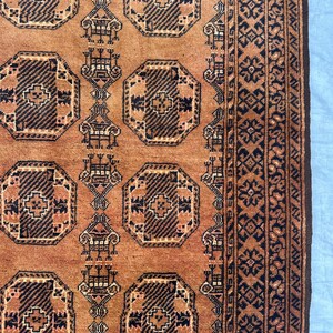 May include: A woven rug featuring a warm terracotta orange background with repeating dark brown octagonal geometric motifs. The design includes vertical columns of stylized symbols and a wide border with intricate dark brown floral patterns.