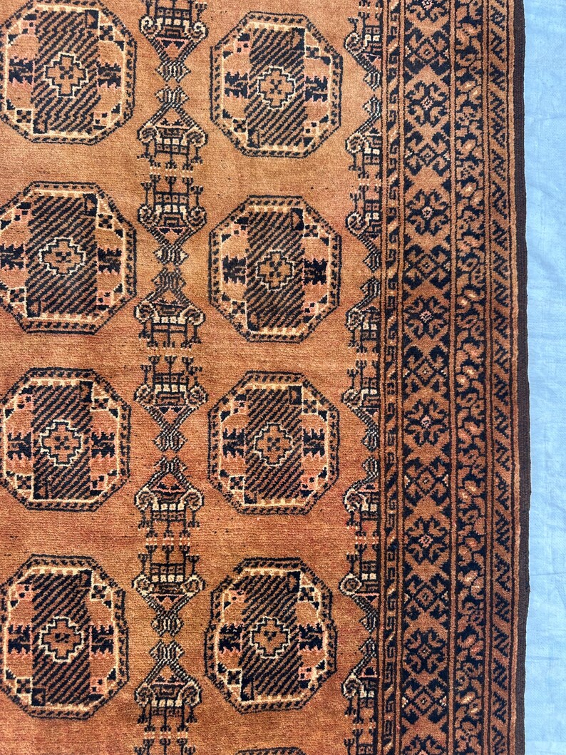 May include: A woven rug featuring a warm terracotta orange background with repeating dark brown octagonal geometric motifs. The design includes vertical columns of stylized symbols and a wide border with intricate dark brown floral patterns.