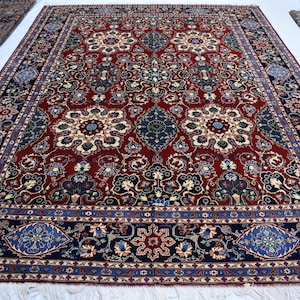 K&ouml;nnte beinhalten: Ein rechteckiger Teppich mit einem tiefroten Grund, verziert mit komplexen Blumen- und Medaillonmustern in Marineblau, Creme und Hellblau. Die Bord&uuml;re zeigt ein sich wiederholendes geometrisches und florales Design in Dunkelblau mit wei&szlig;en Fransen.