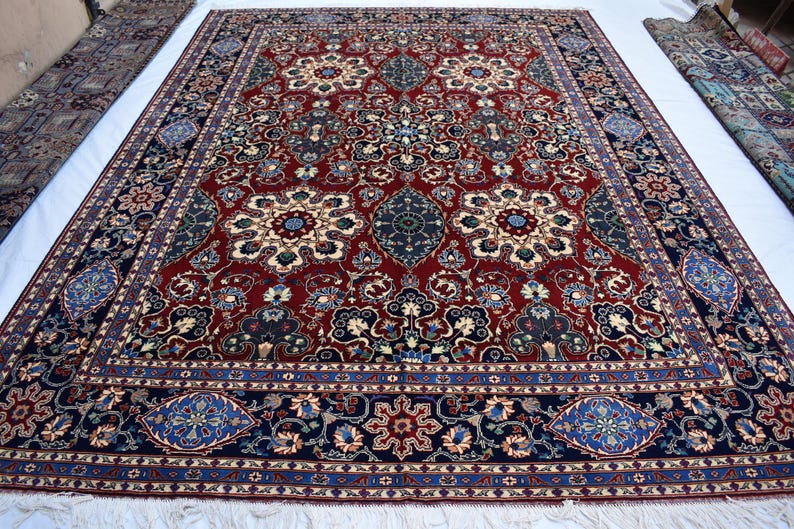 K&ouml;nnte beinhalten: Ein rechteckiger Teppich mit einem tiefroten Grund, verziert mit komplexen Blumen- und Medaillonmustern in Marineblau, Creme und Hellblau. Die Bord&uuml;re zeigt ein sich wiederholendes geometrisches und florales Design in Dunkelblau mit wei&szlig;en Fransen.