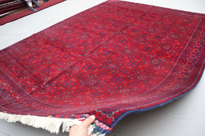 May include: A red and blue patterned rug with a white fringe. The rug is folded in half and is being held by a hand.