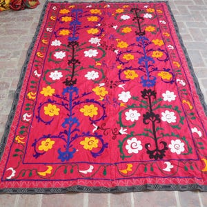 K&ouml;nnte beinhalten: Ein leuchtend rotes Textil mit einem detaillierten Blumenmuster. Das Design zeigt gelbe, wei&szlig;e und blaue Blumen mit schwarzen und gr&uuml;nen Akzenten. Der Rand ist dunkelgrau mit einem geometrischen Muster.