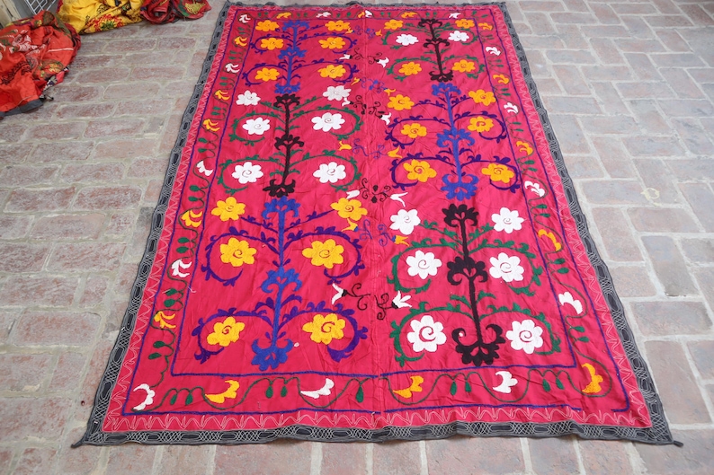 K&ouml;nnte beinhalten: Ein leuchtend rotes Textil mit einem detaillierten Blumenmuster. Das Design zeigt gelbe, wei&szlig;e und blaue Blumen mit schwarzen und gr&uuml;nen Akzenten. Der Rand ist dunkelgrau mit einem geometrischen Muster.
