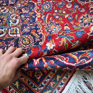 Puede incluir: Una alfombra rectangular con un patr&oacute;n denso y complejo de flores y cachemir en tonos vibrantes de rojo, azul profundo, crema y oro. La alfombra tiene una textura de pelo grueso e incluye un borde con flecos blancos en la parte inferior.