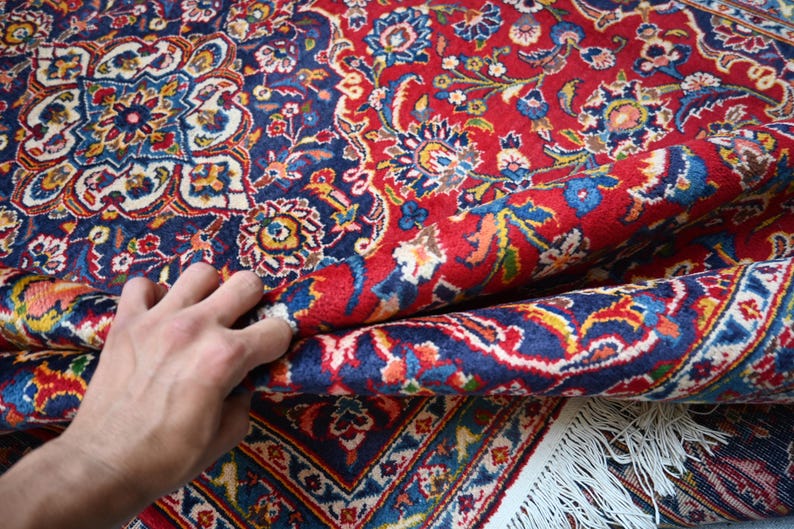 Puede incluir: Una alfombra rectangular con un patr&oacute;n denso y complejo de flores y cachemir en tonos vibrantes de rojo, azul profundo, crema y oro. La alfombra tiene una textura de pelo grueso e incluye un borde con flecos blancos en la parte inferior.
