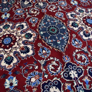 K&ouml;nnte beinhalten: Ein tiefroter Teppich mit einem dichten, komplizierten Muster aus gro&szlig;en, symmetrischen Blumenmedaillons in Marineblau, Creme und Hellblau, durchsetzt mit kleineren botanischen Motiven und einem zentralen, l&auml;nglichen, dunkelblauen blattf&ouml;rmigen Element.