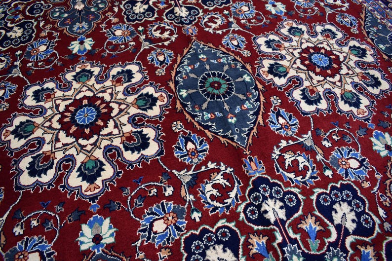 K&ouml;nnte beinhalten: Ein tiefroter Teppich mit einem dichten, komplizierten Muster aus gro&szlig;en, symmetrischen Blumenmedaillons in Marineblau, Creme und Hellblau, durchsetzt mit kleineren botanischen Motiven und einem zentralen, l&auml;nglichen, dunkelblauen blattf&ouml;rmigen Element.