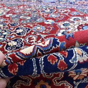K&ouml;nnte beinhalten: Ein dicker Teppich mit tiefrotem Hintergrund und komplizierten Blumen- und geometrischen Mustern in Marineblau, Creme und Hellblau. Der Teppich ist umgeschlagen, um die dichte Florstruktur und das detaillierte Webdesign auf der Oberseite zu zeigen.