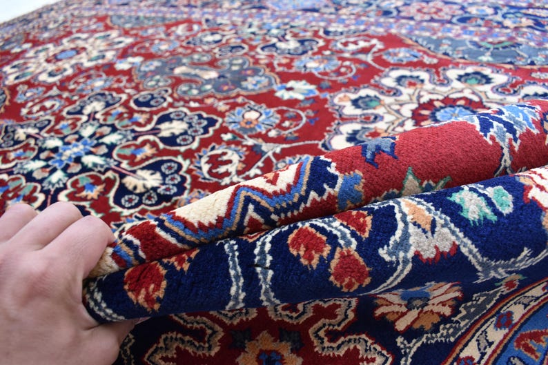 K&ouml;nnte beinhalten: Ein dicker Teppich mit tiefrotem Hintergrund und komplizierten Blumen- und geometrischen Mustern in Marineblau, Creme und Hellblau. Der Teppich ist umgeschlagen, um die dichte Florstruktur und das detaillierte Webdesign auf der Oberseite zu zeigen.