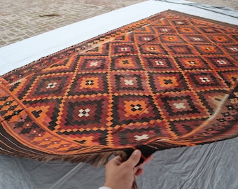 May include: A traditional handwoven rug with a geometric pattern in shades of orange, brown, and black. The rug has a fringe border and is folded in half.