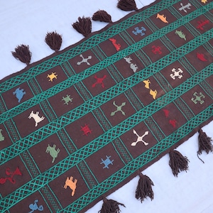 Puede incluir: Un tapete corredor marrón con patrones geométricos verdes y blancos y diseños de animales bordados de colores. La alfombra tiene borlas en cada extremo.