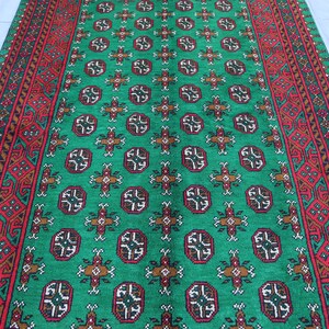 K&ouml;nnte beinhalten: Ein gr&uuml;ner und roter Teppich mit geometrischem Muster. Der Teppich hat einen zentralen gr&uuml;nen Bereich mit einem sich wiederholenden Muster aus kleinen, komplizierten Designs. Die R&auml;nder des Teppichs sind mit einem roten und braunen Muster eingefasst.