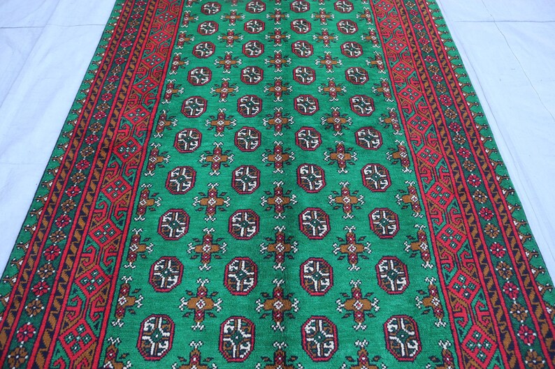 K&ouml;nnte beinhalten: Ein gr&uuml;ner und roter Teppich mit geometrischem Muster. Der Teppich hat einen zentralen gr&uuml;nen Bereich mit einem sich wiederholenden Muster aus kleinen, komplizierten Designs. Die R&auml;nder des Teppichs sind mit einem roten und braunen Muster eingefasst.