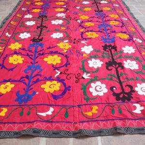 K&ouml;nnte beinhalten: Ein lebendiges, rechteckiges Textil mit rotem Hintergrund. Das Textil weist gestickte Blumenmuster in Gelb, Wei&szlig;, Blau und Schwarz auf. Die R&auml;nder sind mit einem dekorativen Rand versehen.