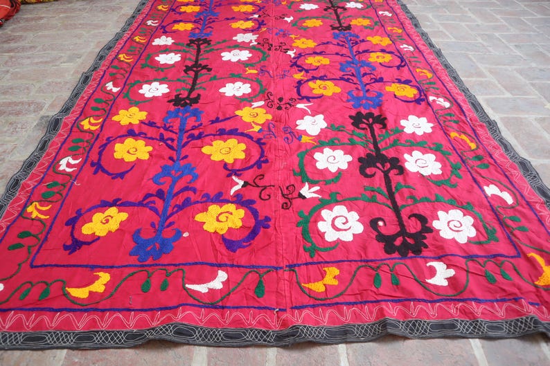 K&ouml;nnte beinhalten: Ein lebendiges, rechteckiges Textil mit rotem Hintergrund. Das Textil weist gestickte Blumenmuster in Gelb, Wei&szlig;, Blau und Schwarz auf. Die R&auml;nder sind mit einem dekorativen Rand versehen.