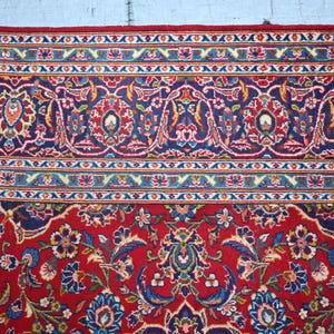 Puede incluir: Una alfombra rectangular con un fondo rojo intenso y complejos patrones florales y de enredaderas en tonos azul, blanco y dorado. El borde muestra una disposici&oacute;n compleja de motivos geom&eacute;tricos y bot&aacute;nicos repetitivos en azul y rojo.