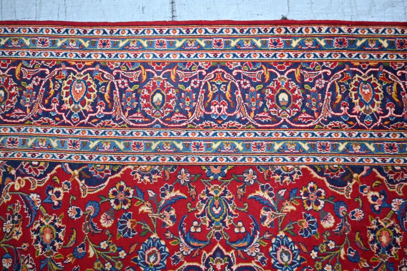 Puede incluir: Una alfombra rectangular con un fondo rojo intenso y complejos patrones florales y de enredaderas en tonos azul, blanco y dorado. El borde muestra una disposici&oacute;n compleja de motivos geom&eacute;tricos y bot&aacute;nicos repetitivos en azul y rojo.
