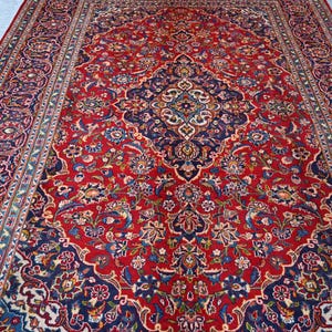Puede incluir: Una alfombra rectangular grande con un fondo rojo vibrante y un medall&oacute;n central azul marino. La superficie est&aacute; cubierta con intrincados patrones florales y de enredaderas en tonos azul claro, crema y dorado, enmarcada por un borde decorativo.