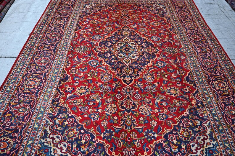 Puede incluir: Una alfombra rectangular grande con un fondo rojo vibrante y un medall&oacute;n central azul marino. La superficie est&aacute; cubierta con intrincados patrones florales y de enredaderas en tonos azul claro, crema y dorado, enmarcada por un borde decorativo.