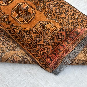 May include: A rectangular area rug featuring a deep orange and black geometric pattern with repeating octagonal motifs. The rug is folded over to reveal the underside, showcasing a dense weave and a short, grey fringe along the edge of the fabric.