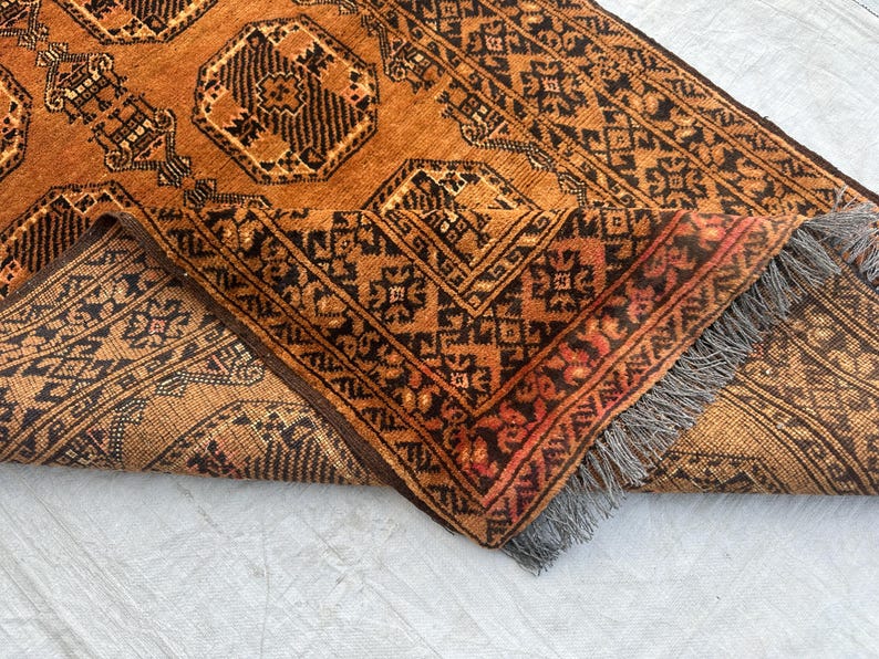 May include: A rectangular area rug featuring a deep orange and black geometric pattern with repeating octagonal motifs. The rug is folded over to reveal the underside, showcasing a dense weave and a short, grey fringe along the edge of the fabric.