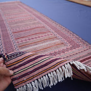 May include: A long, rectangular runner rug featuring a woven pattern of pink, red, and purple horizontal stripes. The rug has a decorative border with geometric motifs and white fringe along the short edge, showing the underside of the fabric.