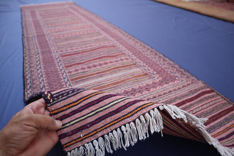 May include: A long, rectangular runner rug featuring a woven pattern of pink, red, and purple horizontal stripes. The rug has a decorative border with geometric motifs and white fringe along the short edge, showing the underside of the fabric.