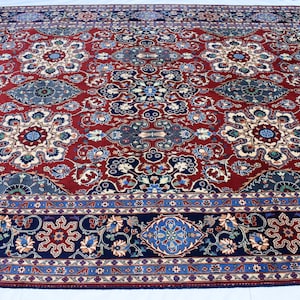 K&ouml;nnte beinhalten: Ein rechteckiger Teppich mit einem tiefroten Grund und einem sich wiederholenden Muster aus floralen Medaillons in Blau-, Creme- und Dunkelmarine-T&ouml;nen. Der Teppich ist von einer breiten, dunkelblauen Bord&uuml;re mit floralen Motiven umrahmt.