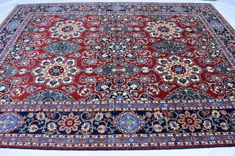 K&ouml;nnte beinhalten: Ein rechteckiger Teppich mit einem tiefroten Grund und einem sich wiederholenden Muster aus floralen Medaillons in Blau-, Creme- und Dunkelmarine-T&ouml;nen. Der Teppich ist von einer breiten, dunkelblauen Bord&uuml;re mit floralen Motiven umrahmt.