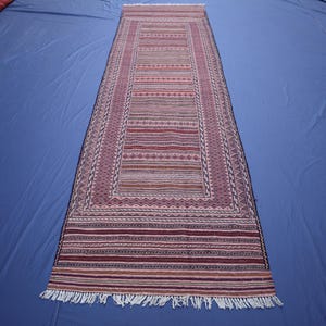 May include: A long, rectangular runner rug featuring a central panel of horizontal stripes in shades of red, pink, and white, surrounded by a geometric border pattern. The rug ends in white fringe at both ends, showcasing a woven textile design.