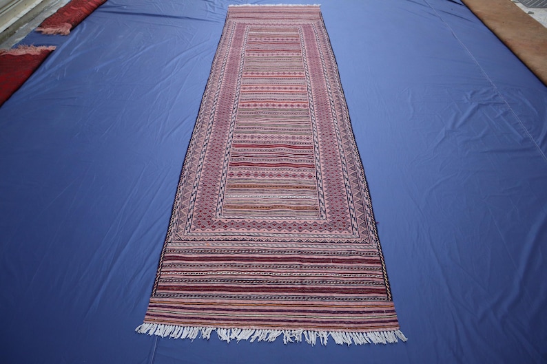 May include: A long, rectangular runner rug featuring a central panel of horizontal stripes in shades of red, pink, and white, surrounded by a geometric border pattern. The rug ends in white fringe at both ends, showcasing a woven textile design.