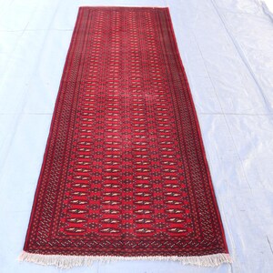 Puede incluir: Una alfombra rectangular roja con un intrincado patrón geométrico. La alfombra presenta un diseño repetitivo de formas de diamante en un tono más oscuro de rojo y marrón, con una franja blanca en ambos extremos. La alfombra probablemente está hecha de material tejido.