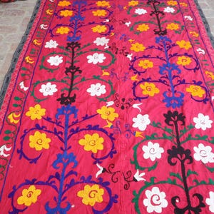 K&ouml;nnte beinhalten: Ein rechteckiger Textil mit pinkfarbenem Hintergrund. Das Design zeigt stilisierte Blumenmuster in Gelb, Wei&szlig;, Blau und Gr&uuml;n. Die R&auml;nder sind mit einem schwarz-grauen Muster eingefasst. Dieses dekorative Textil eignet sich f&uuml;r die Inneneinrichtung.